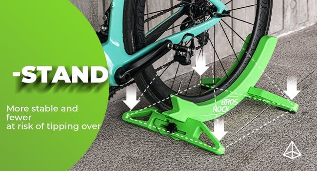 Green bicycle stand with cross-shaped base. Arrows point to its corners, indicating stability. Text highlights reduced tipping risk. Teal bicycle tire visible.